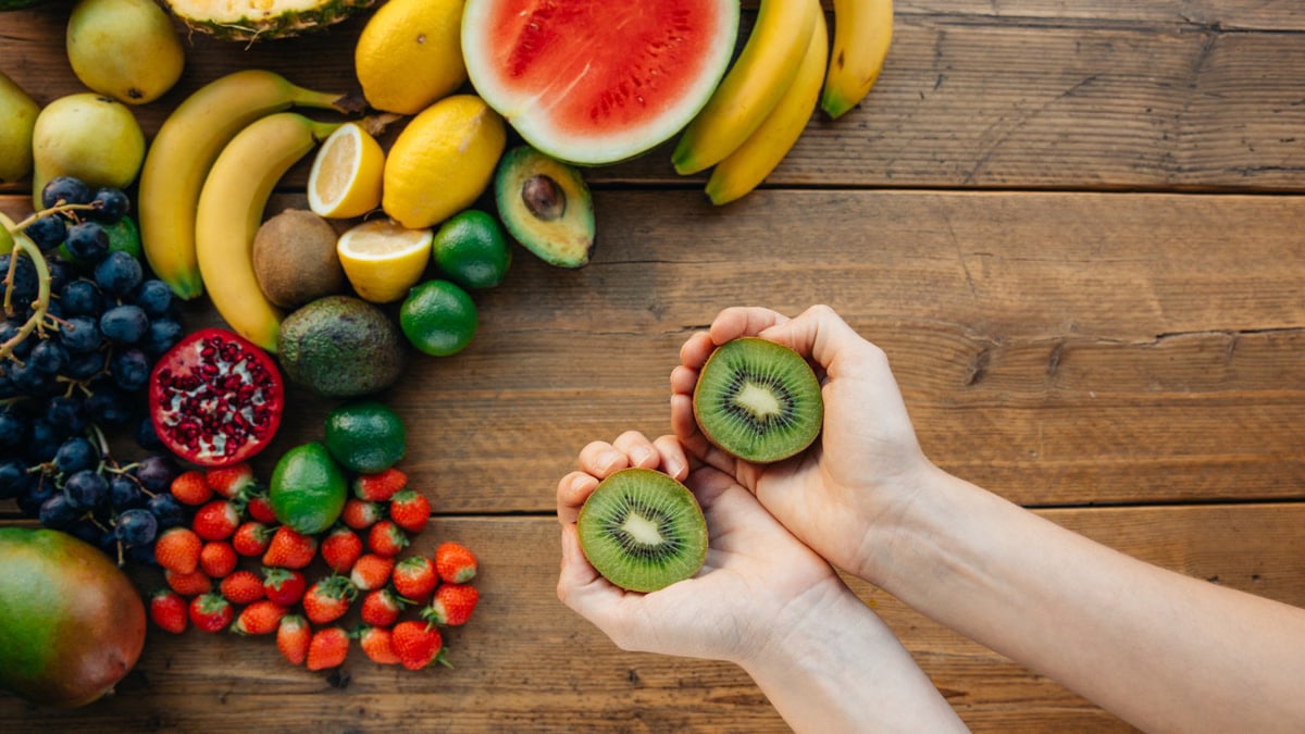 Hands holding kiwis with a variety of fresh fruits on a wooden table.